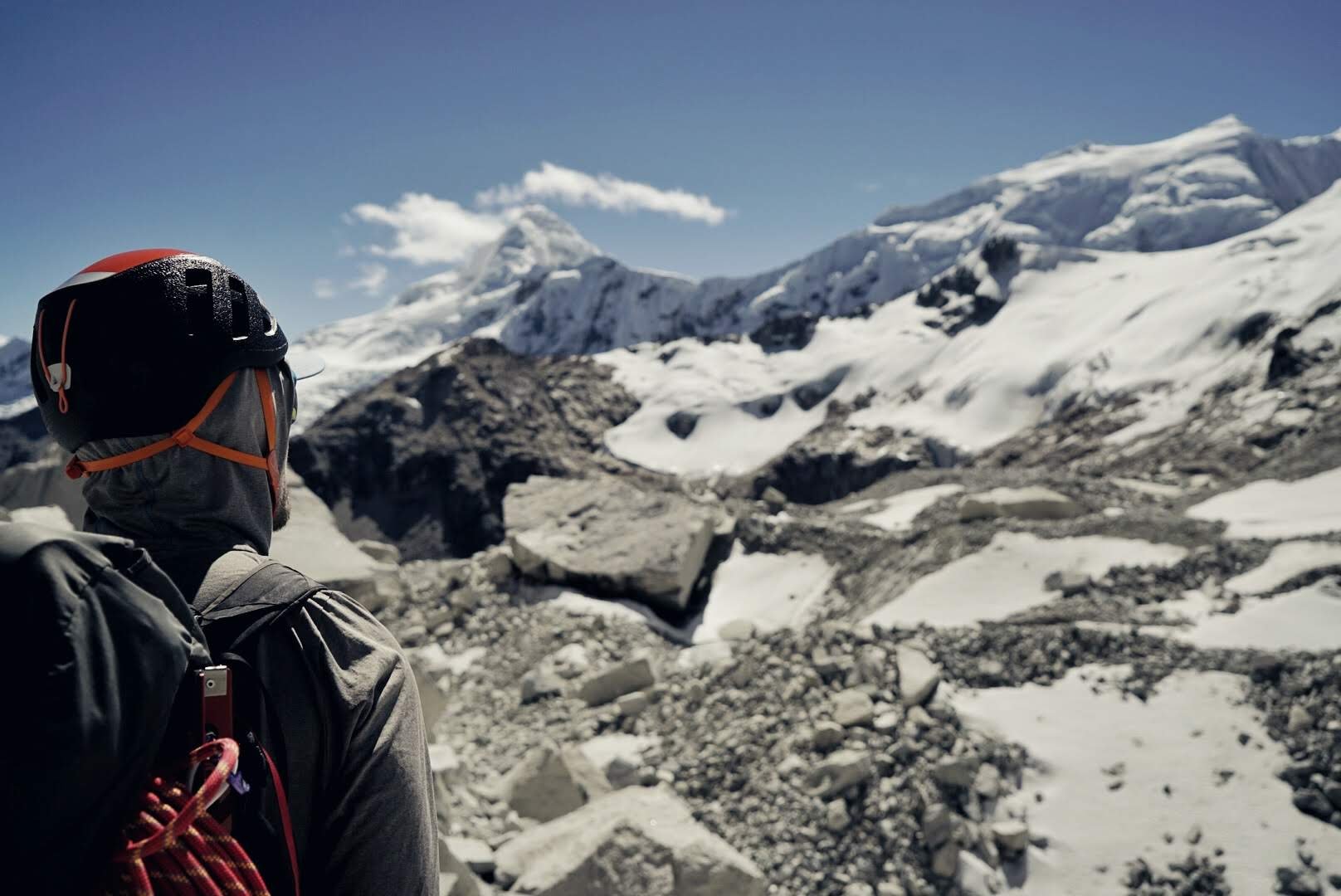 Person wearing a helmet and outdoor gear overlooking a snow-covered mountain landscape with rocks and glaciers.