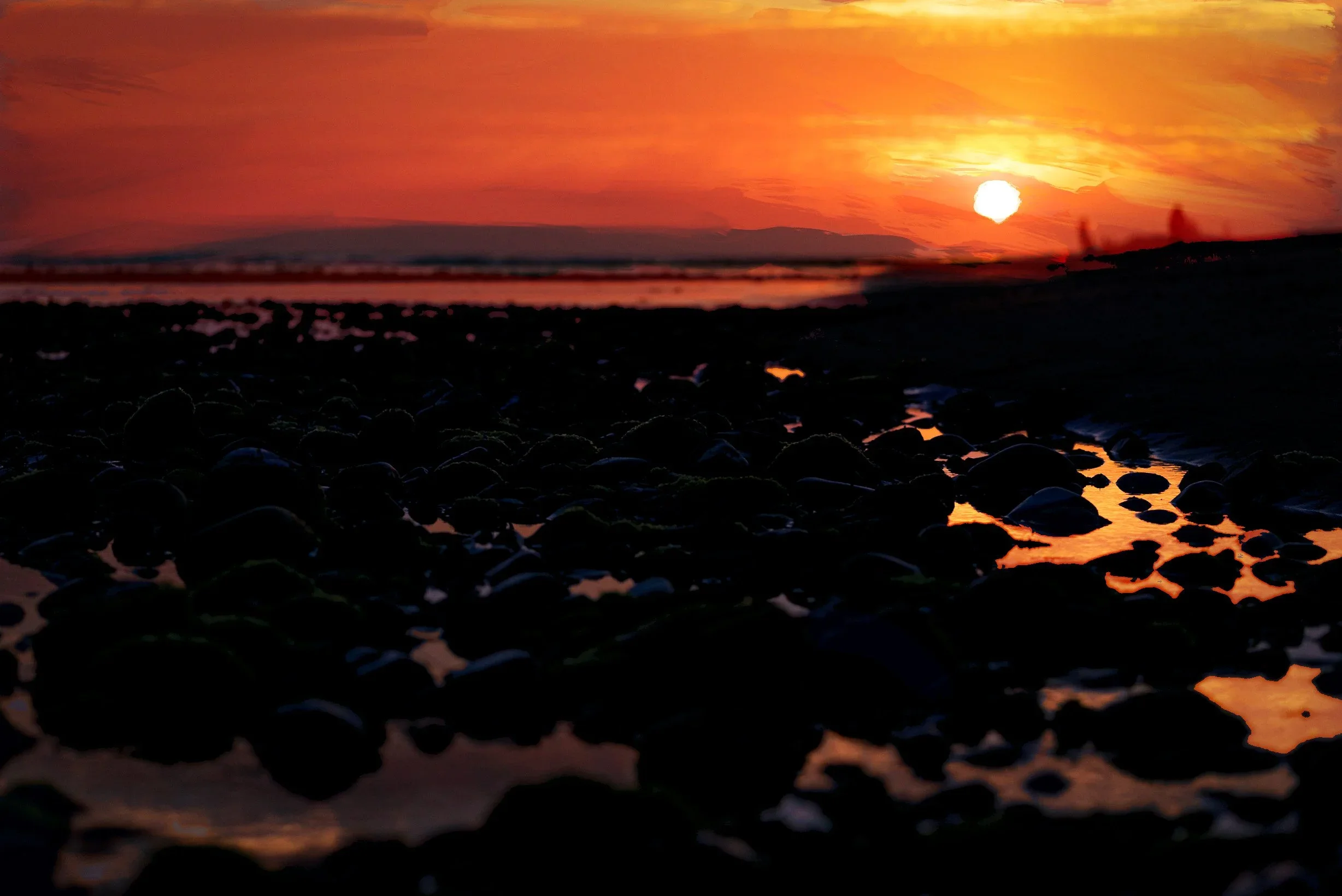 Sunset over a beach with rocks in the foreground and two silhouettes walking along the shoreline.
