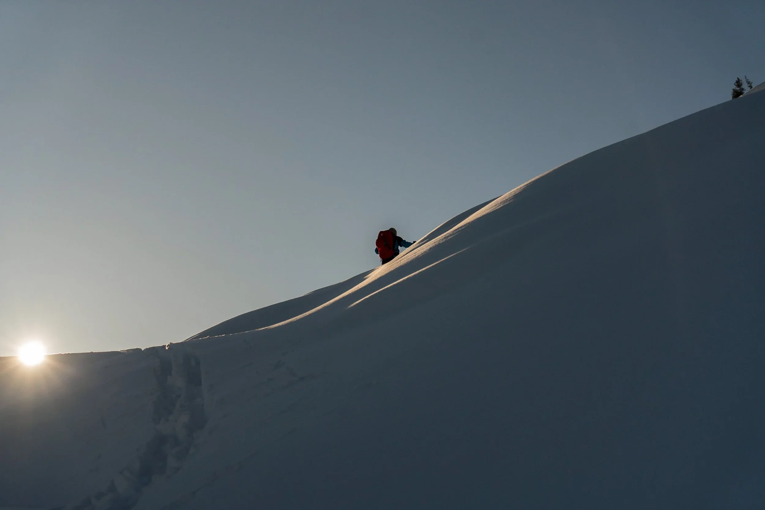 Snow-covered mountain slope with a skier in red gear ascending, with the sun low on the horizon, and a few trees visible at the top of the slope.