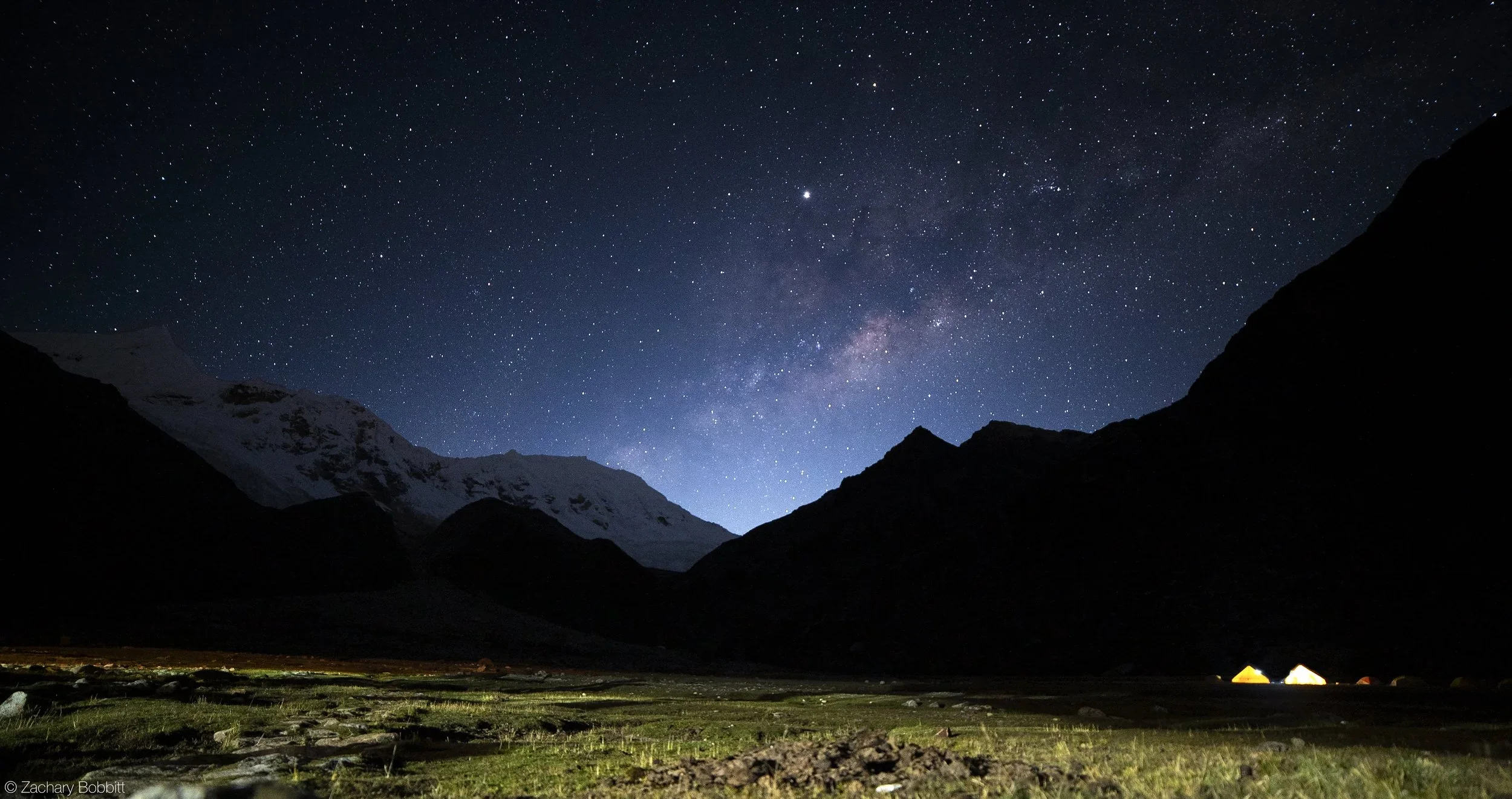 Nighttime mountain landscape with a star-filled sky, including the Milky Way galaxy, behind silhouetted mountain peaks and a small camp with tents illuminated by a yellow light.