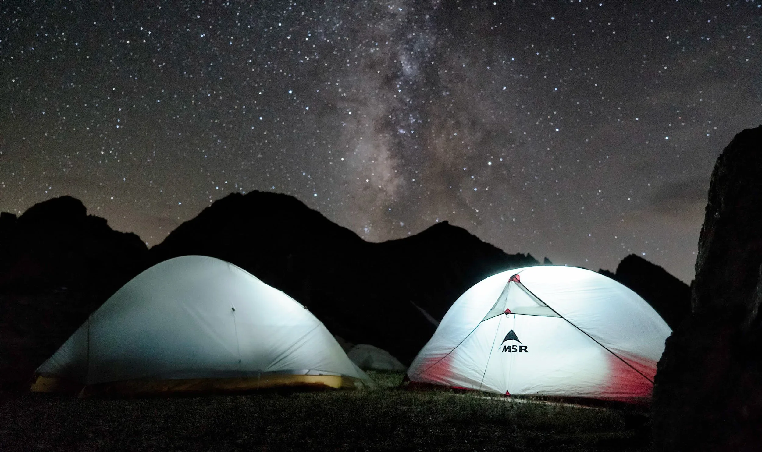 Night scene with a star-filled sky and the Milky Way galaxy visible above two tents set up in a rocky outdoor area.