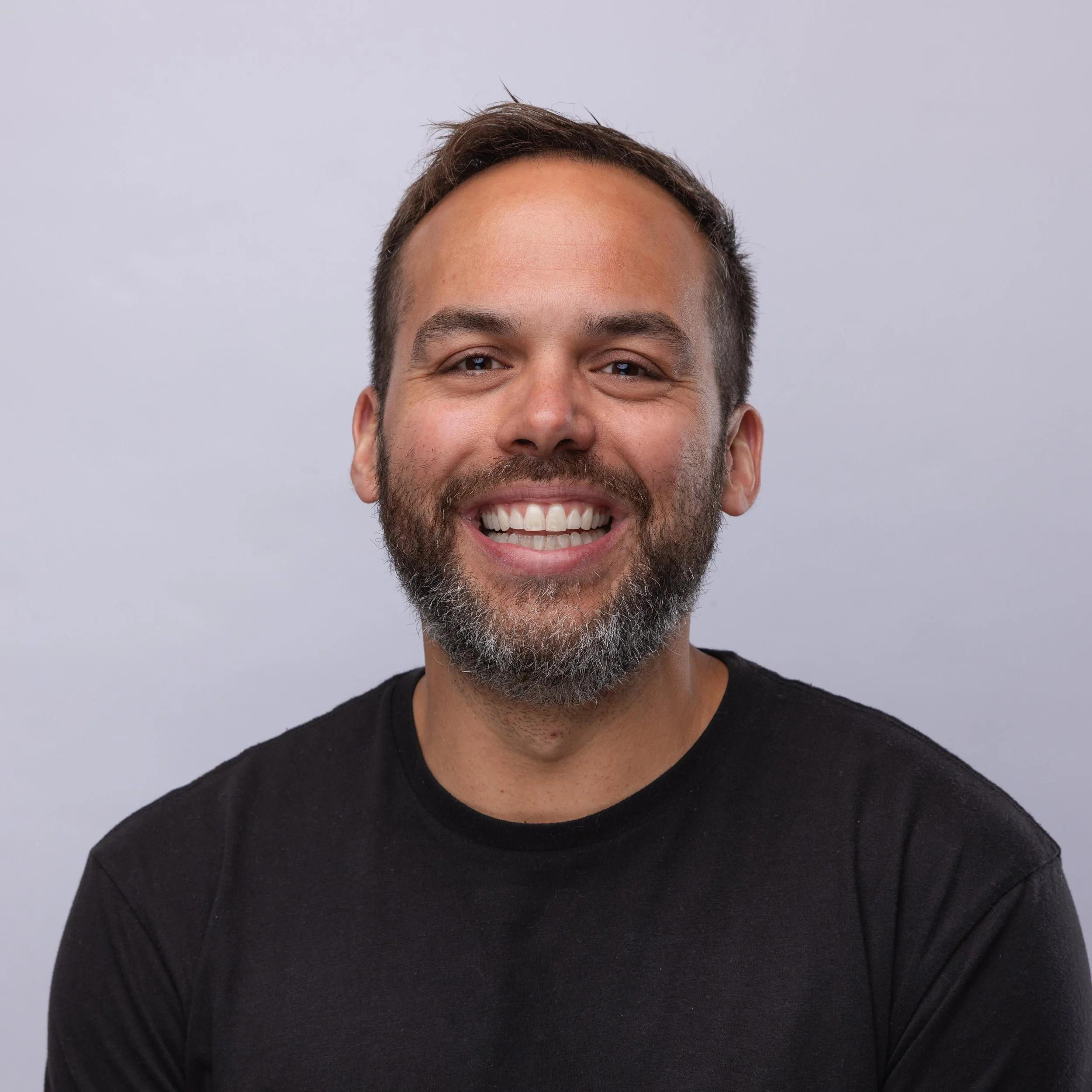 Close-up of a smiling man with short brown hair, light skin, a beard, wearing a black shirt, against a plain light gray background.