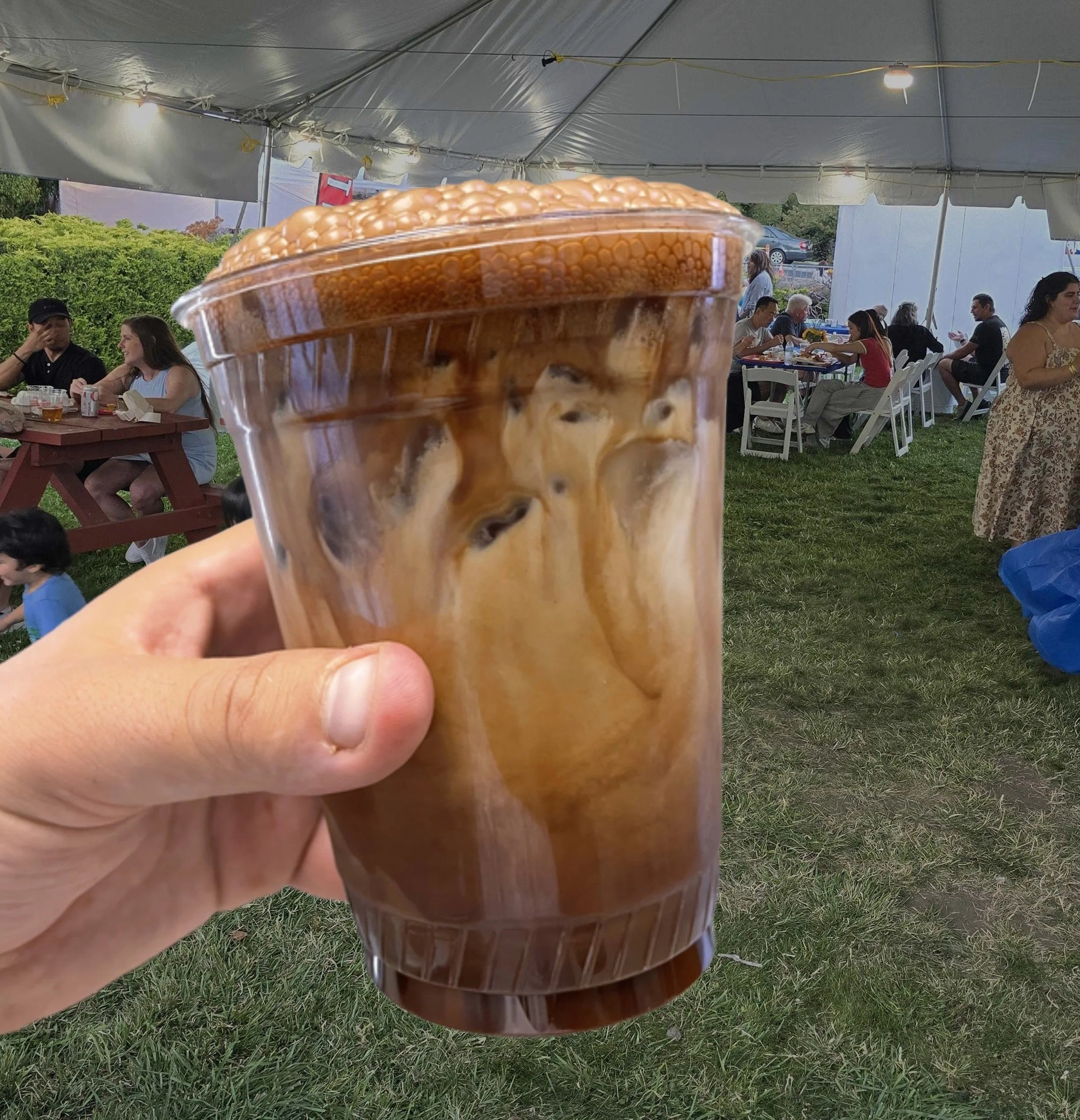 A hand holding a plastic cup of Greek frappe, iced coffee, at a large outdoor tent with people sitting at tables at the Belmont Greek Festival.