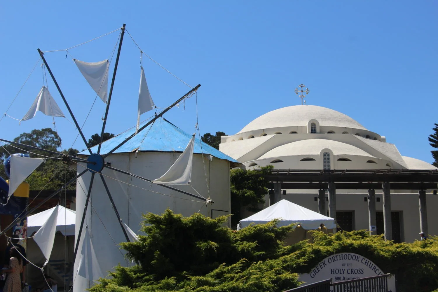 A white church with a large dome and a cross on top, surrounded by trees and tents, with a blue sky in the background.