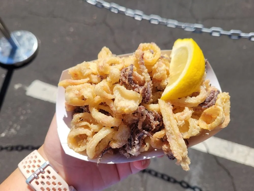 A hand holding a paper boat filled with fried kalamari and a lemon wedge at the Belmont Greek Festival
