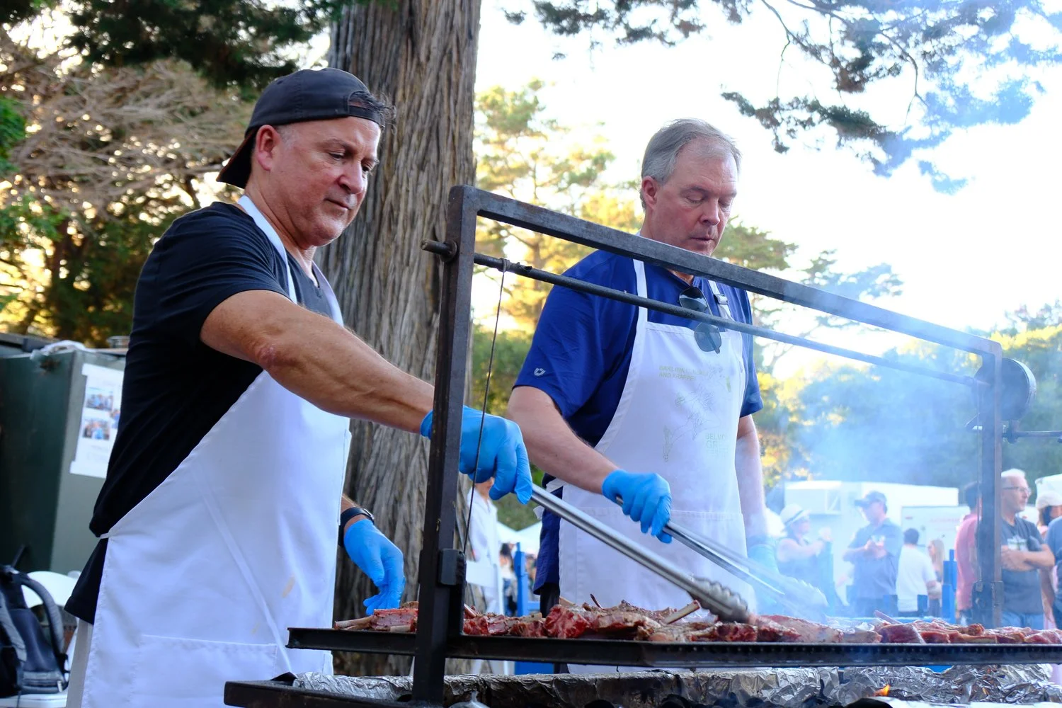 Two men wearing white aprons and blue gloves cook lamb chops on a large outdoor grill at the festival during daytime.