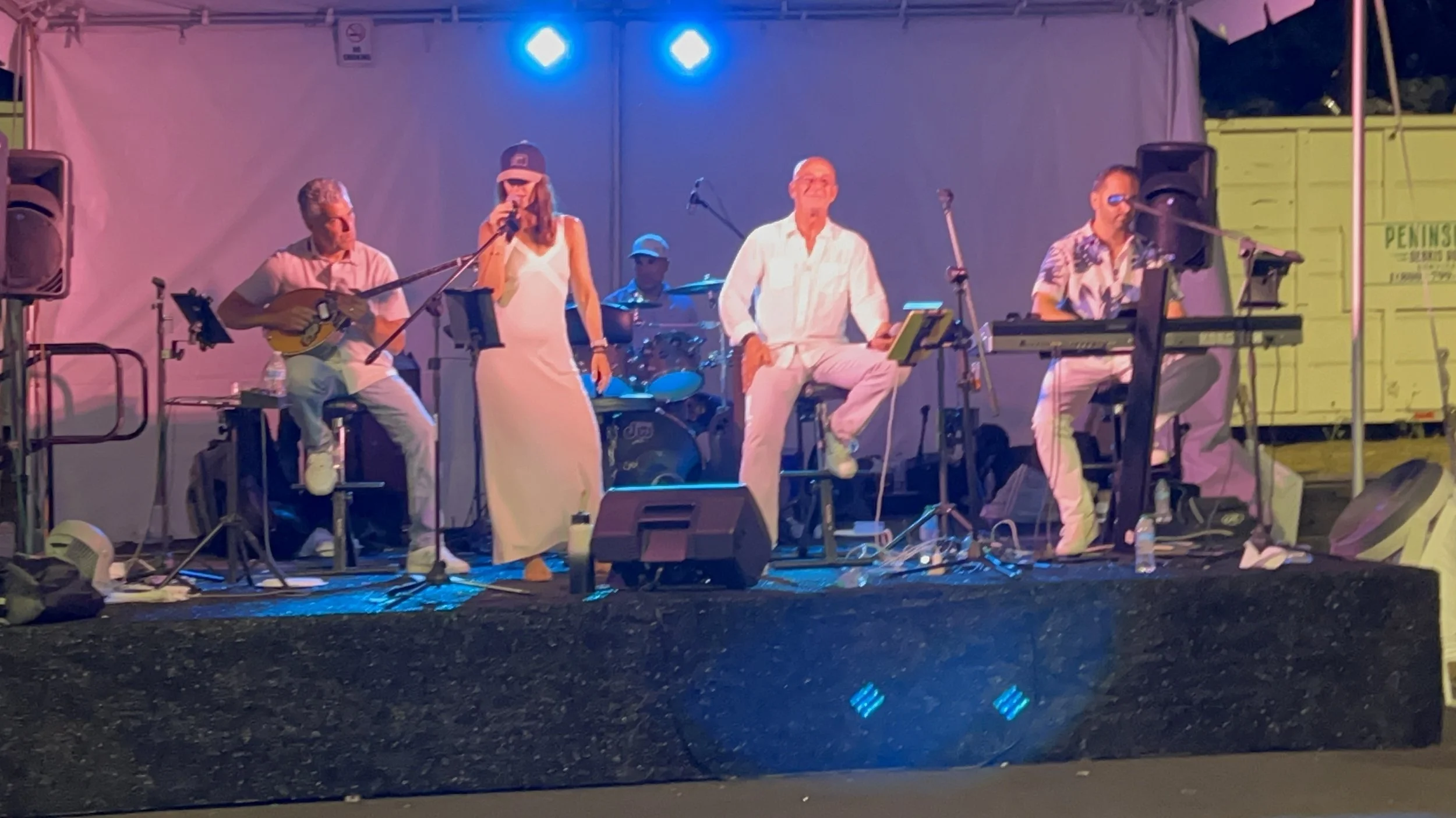 Mythos Band performing on stage with four musicians, one playing Greek bouzouki, one singing, one on drums, and one on keyboard, with colorful stage lighting.