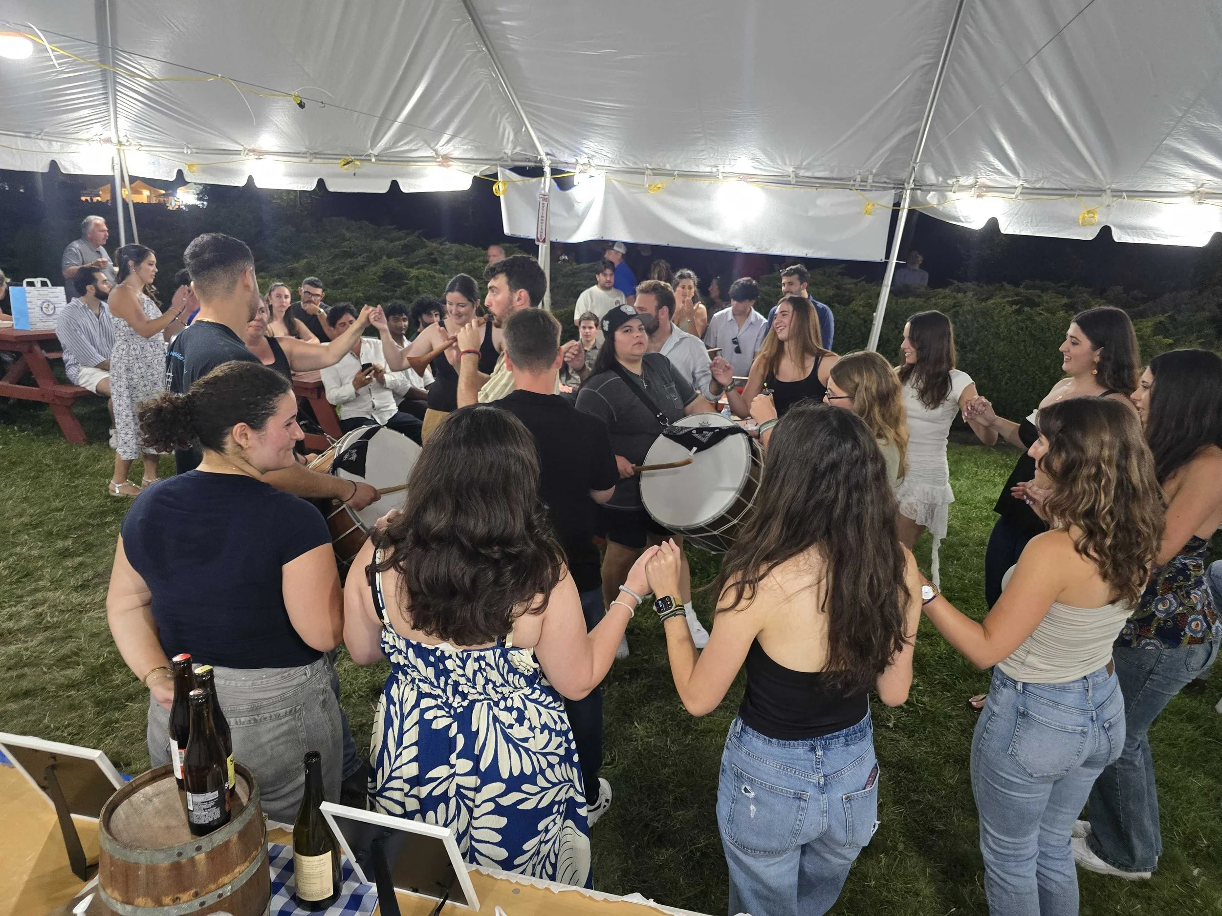 A large group of young people Greek folk dancing and socializing under a white tent at the Mykonos Beer Garden at night. Some are holding beverages and others are clasping hands in a dance circle.
