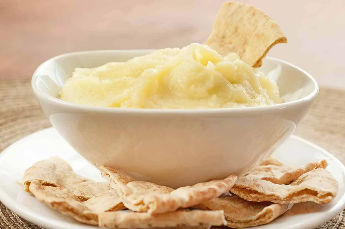 A bowl of Greek skordalia dip with a pita triangle on top, served on a plate with additional pita around it.