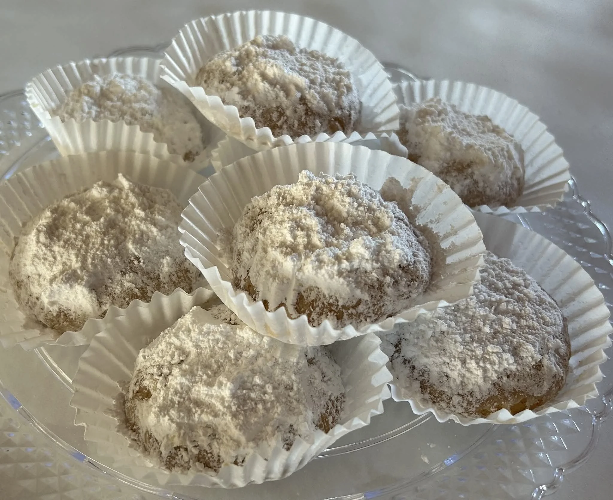 A stack of Greek kouroumbiedes covered in powdered sugar and placed in white ridged paper cups, arranged on a clear plastic tray.