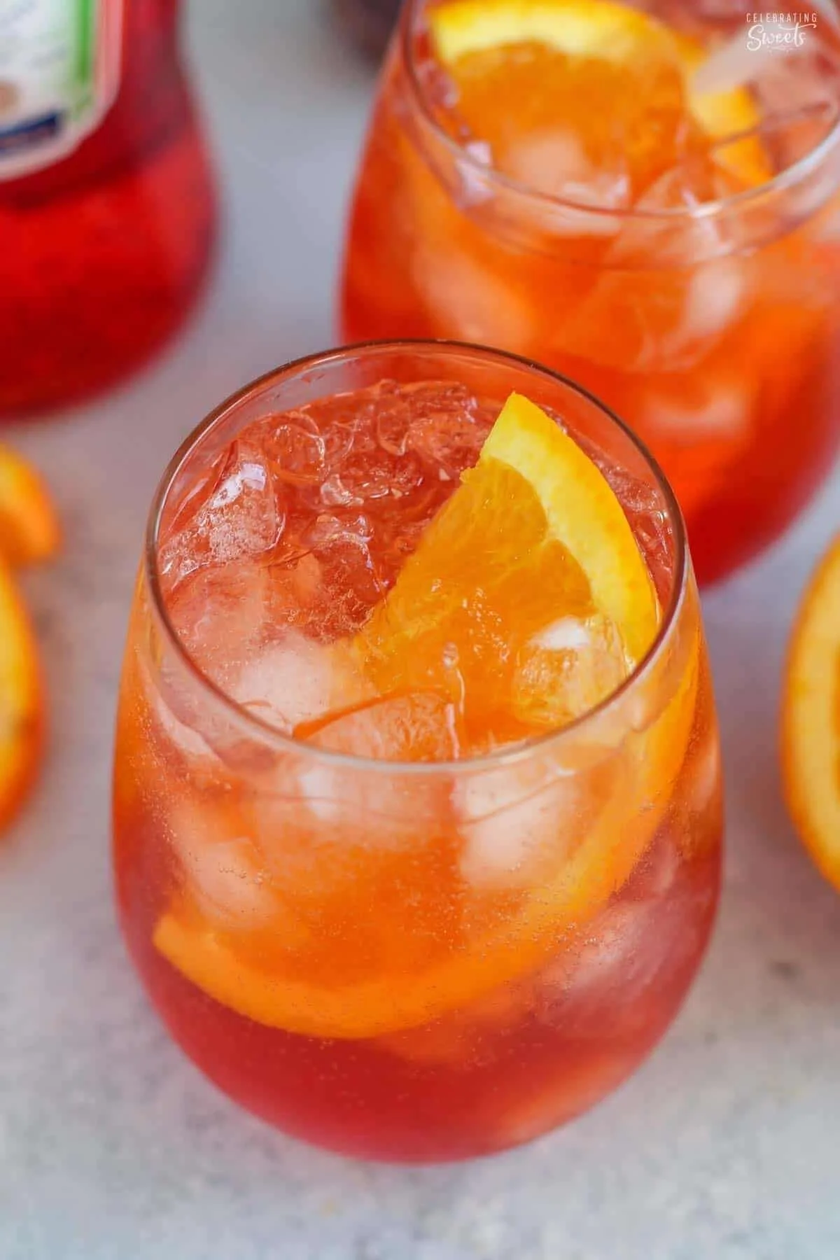 A glass of aperol spritz, an orange cocktail with ice and a wedge of orange on top. Additional glasses and a pitcher of the same drink are in the background.