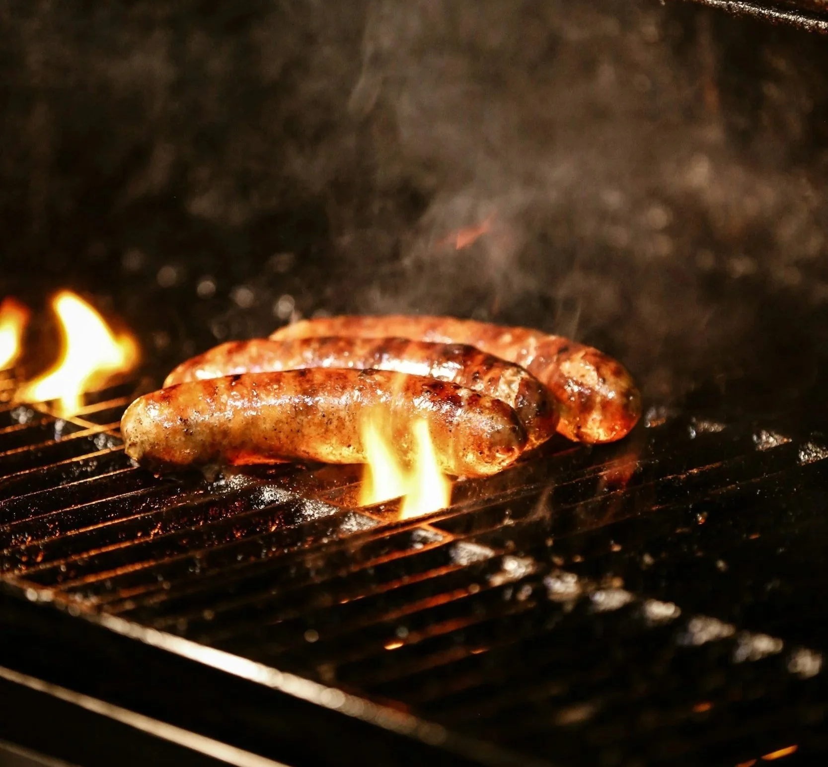 Three loukanika, Greek sausages, cooking on a barbecue grill over open flames, with smoke rising.