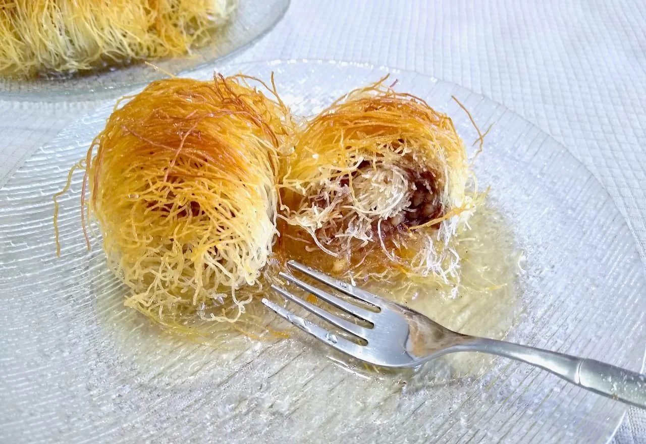 Close-up of two pieces of kataifi, a Greek dessert with shredded phyllo dough, on a clear glass plate, one of which is cut open to reveal a nut filling, with syrup dripping on the plate and a fork nearby.