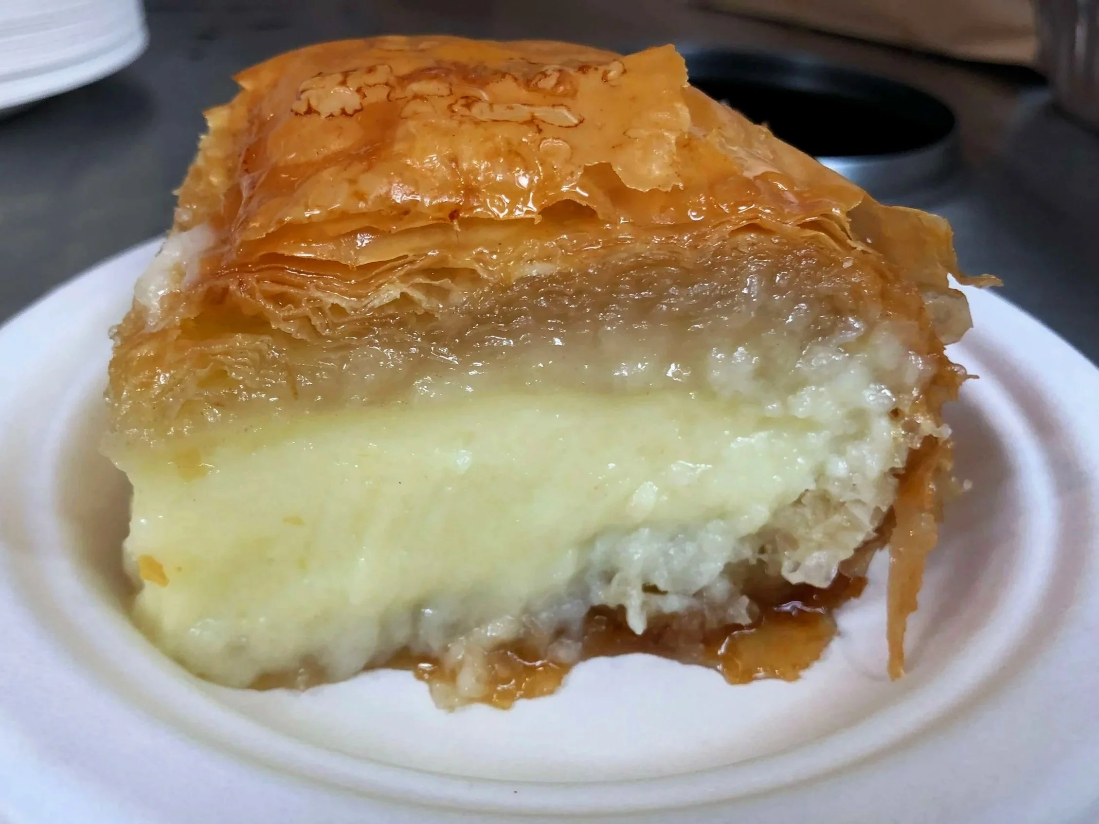 A piece of galactoboureko on a white plate showing flaky layers of phyllo dough, with a sticky syrup glaze on top.