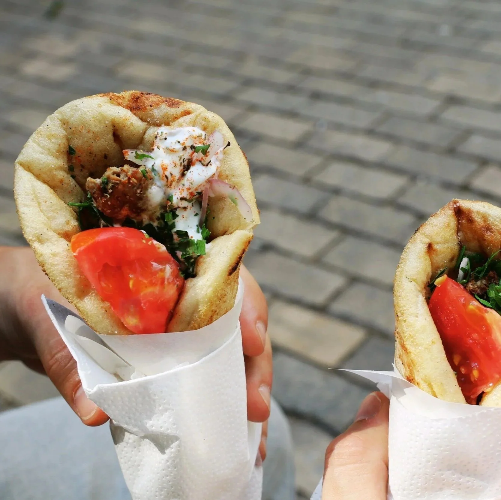 Two hands holding up Greek gyros with lamb, tomato, and tzatziki