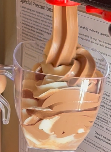 A clear plastic cup with chocolate and vanilla soft serve ice cream being drizzled in from an ice cream machine