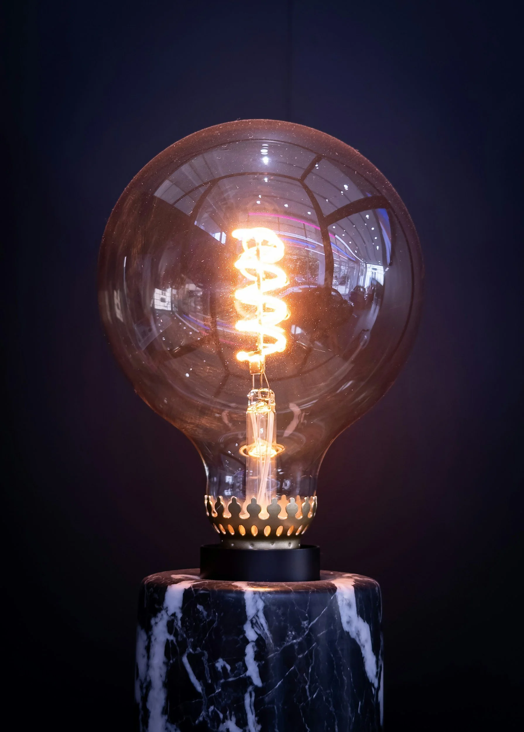 A close-up of an illuminated Edison-style light bulb with a glowing filament, mounted on a black and white marble base, against a dark background.
