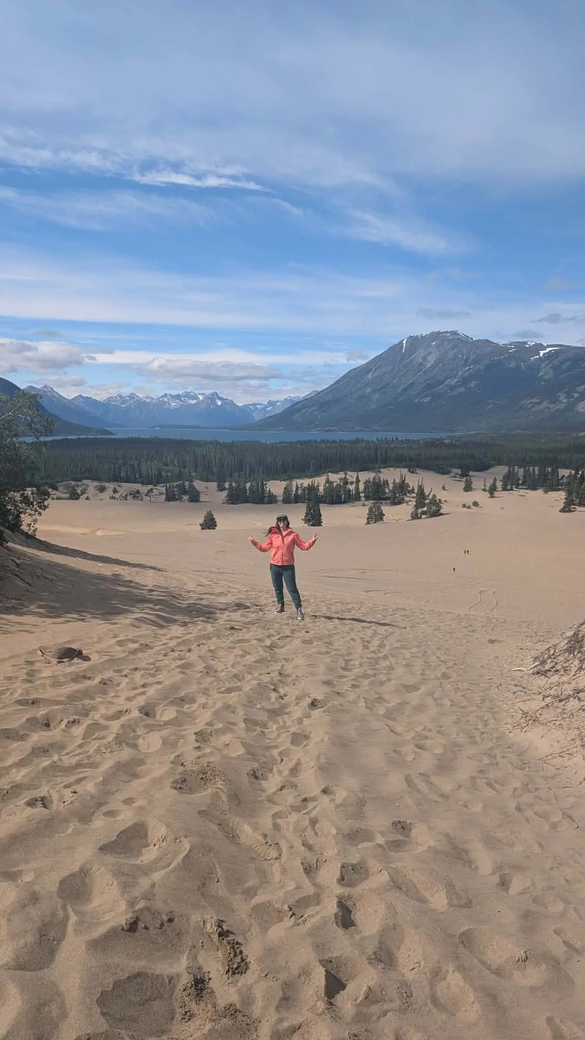 A person in a pink jacket and black pants standing on sandy terrain, posing with arms outstretched in front of a mountain and lake landscape under a partly cloudy sky.