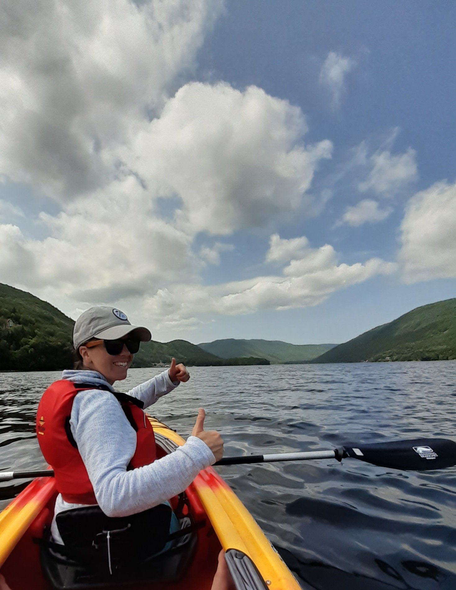 A woman in sunglasses smiling and giving a thumbs-up while kayaking on a lake surrounded by green hills under a partly cloudy sky.