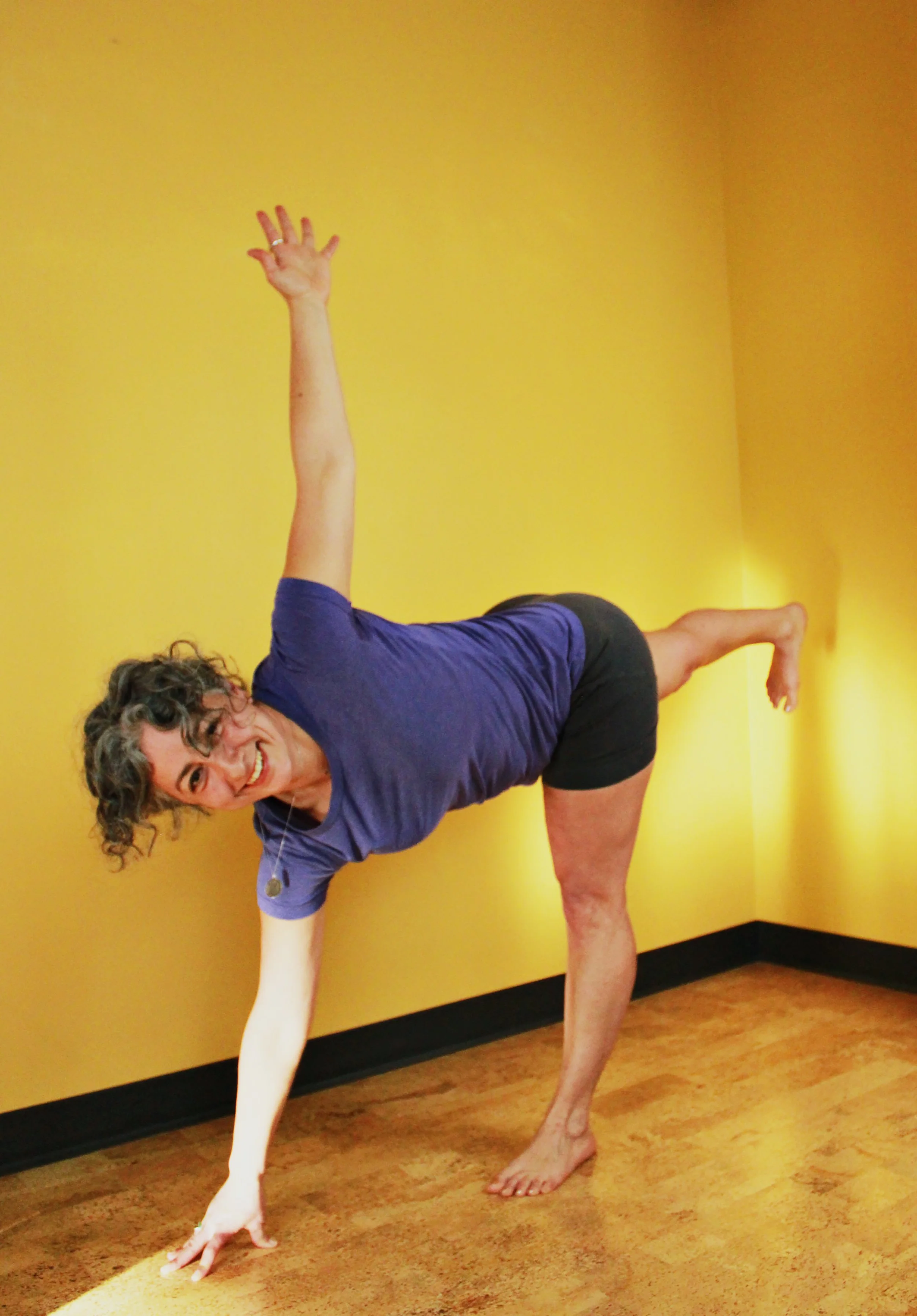A woman with curly hair smiling and wearing a purple t-shirt and black shorts performing a yoga pose with one hand and foot on the floor, one arm extended upward, and the other arm reaching forward, in front of a yellow wall.