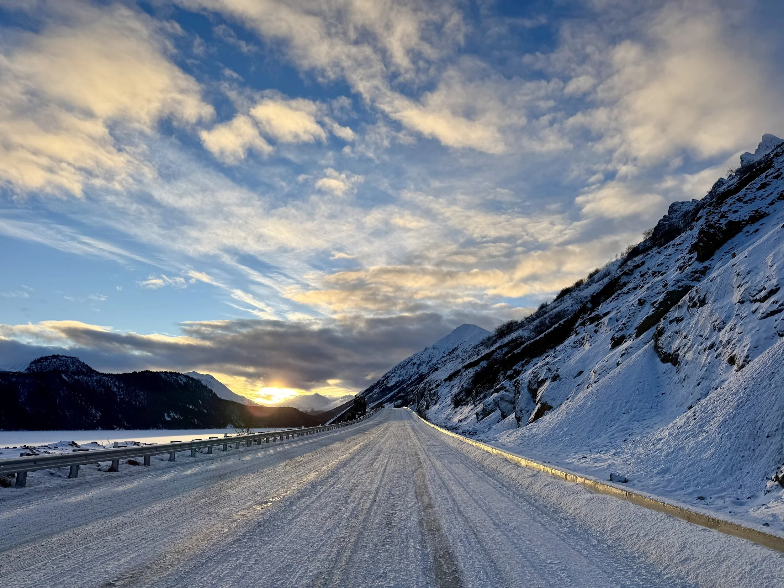 A snow-covered mountain road at sunset with a guardrail on the side, snow on the mountain slopes, and clouds in the sky with the sun low on the horizon.