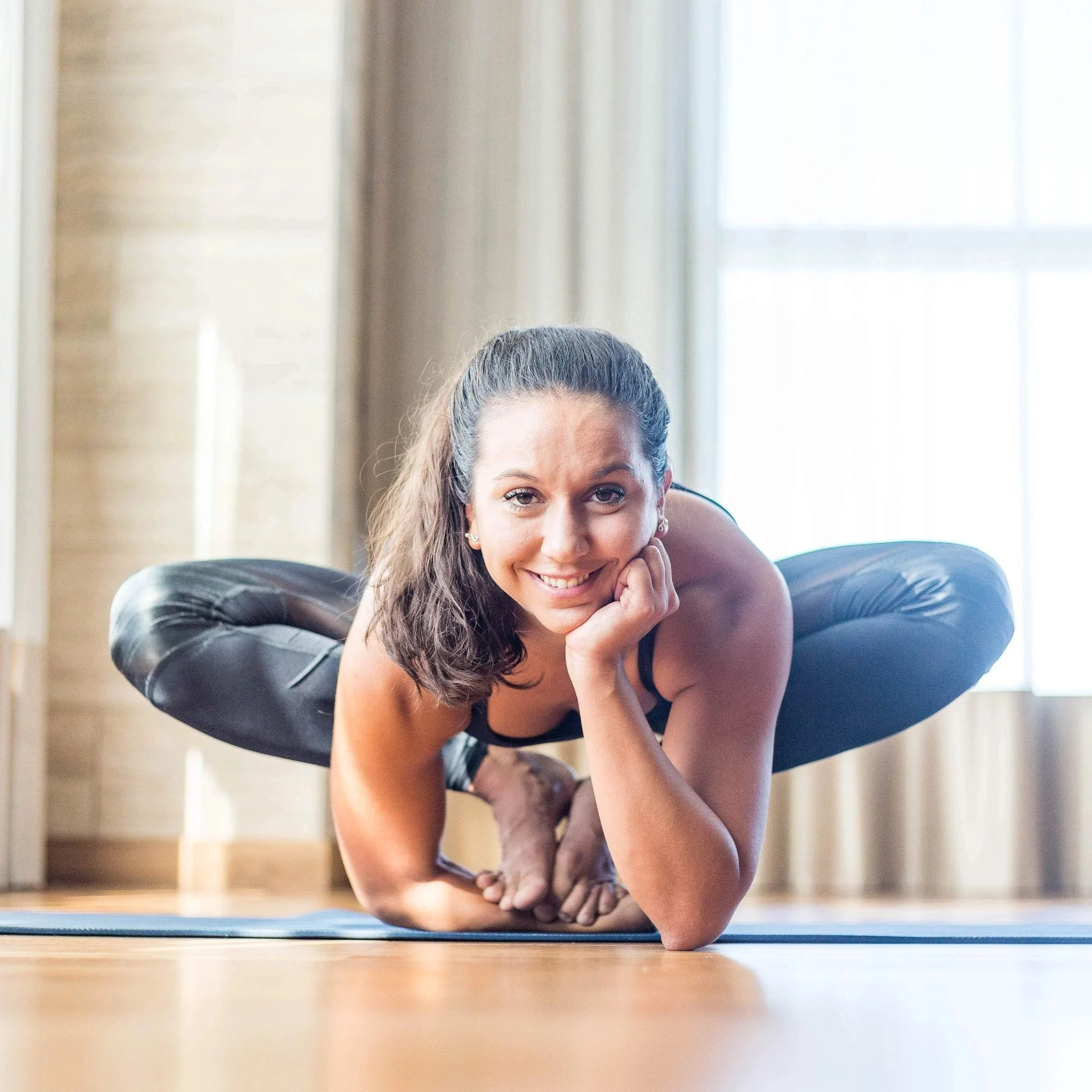 A woman in black workout clothes practicing yoga or stretching on a blue mat in a bright room with large windows.