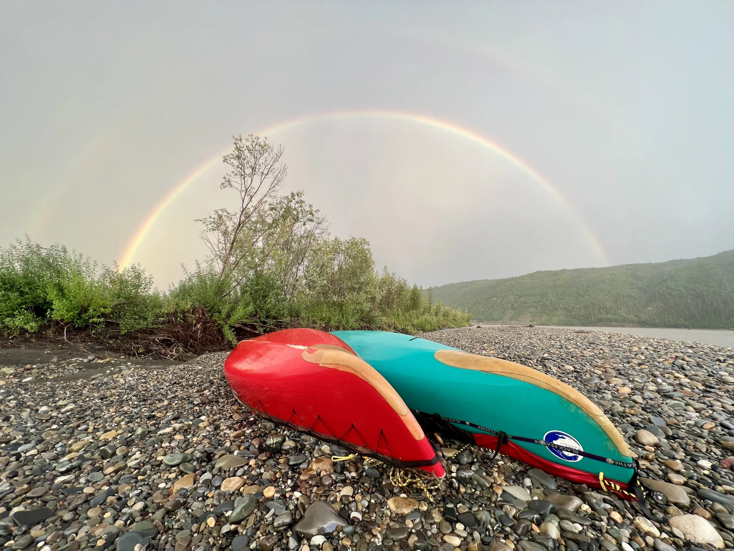 Colorful kayaks lying on a pebble beach with a rainbow in the sky and a tree in the background.