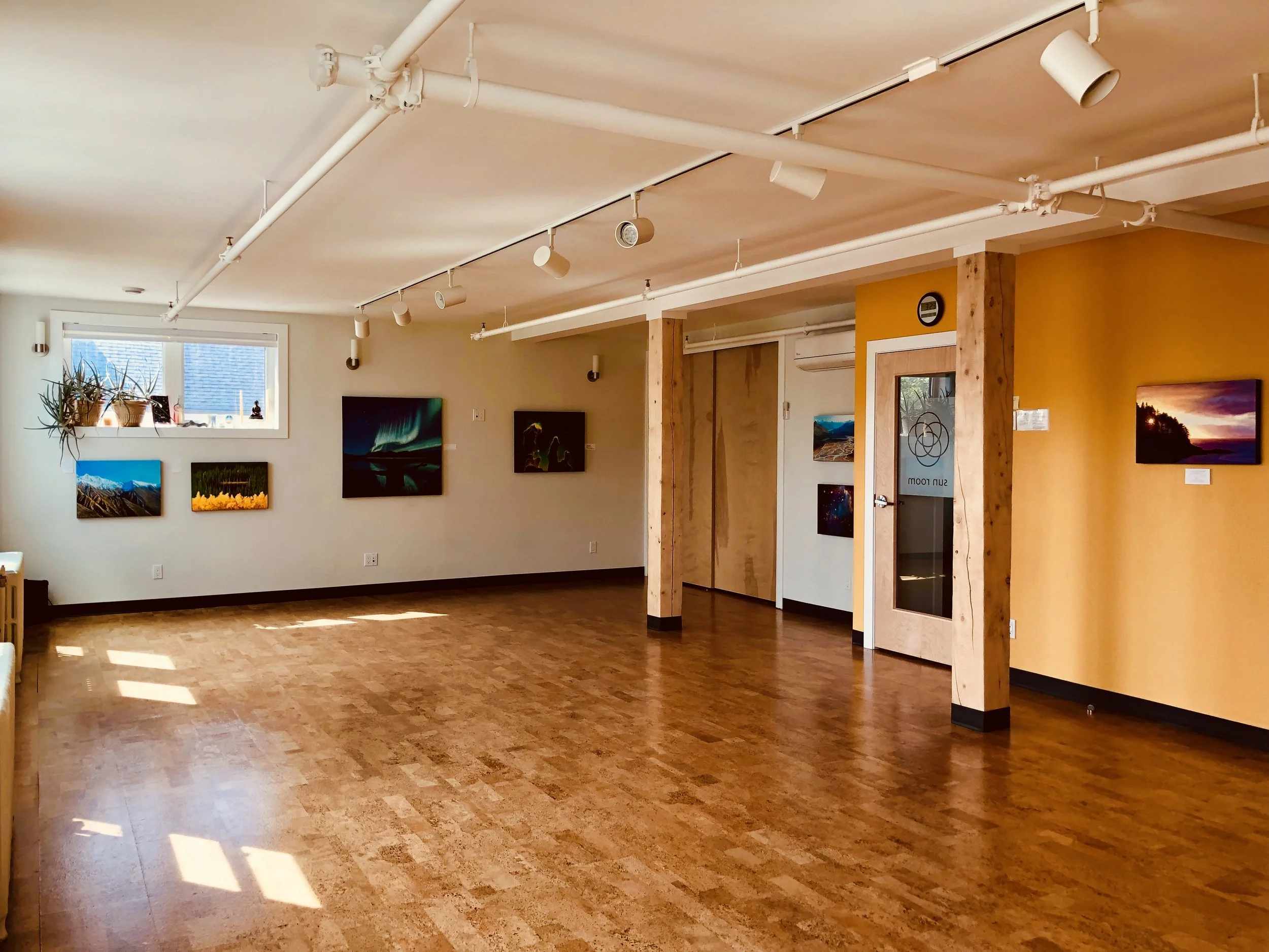An art gallery interior with framed landscape photographs hanging on white and yellow walls, wooden beams, track lighting, and wood flooring.