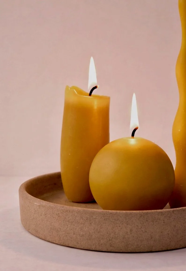 Handmade pure beeswax candles, one round ball candle and one spindle, lit on a ceramic dish.