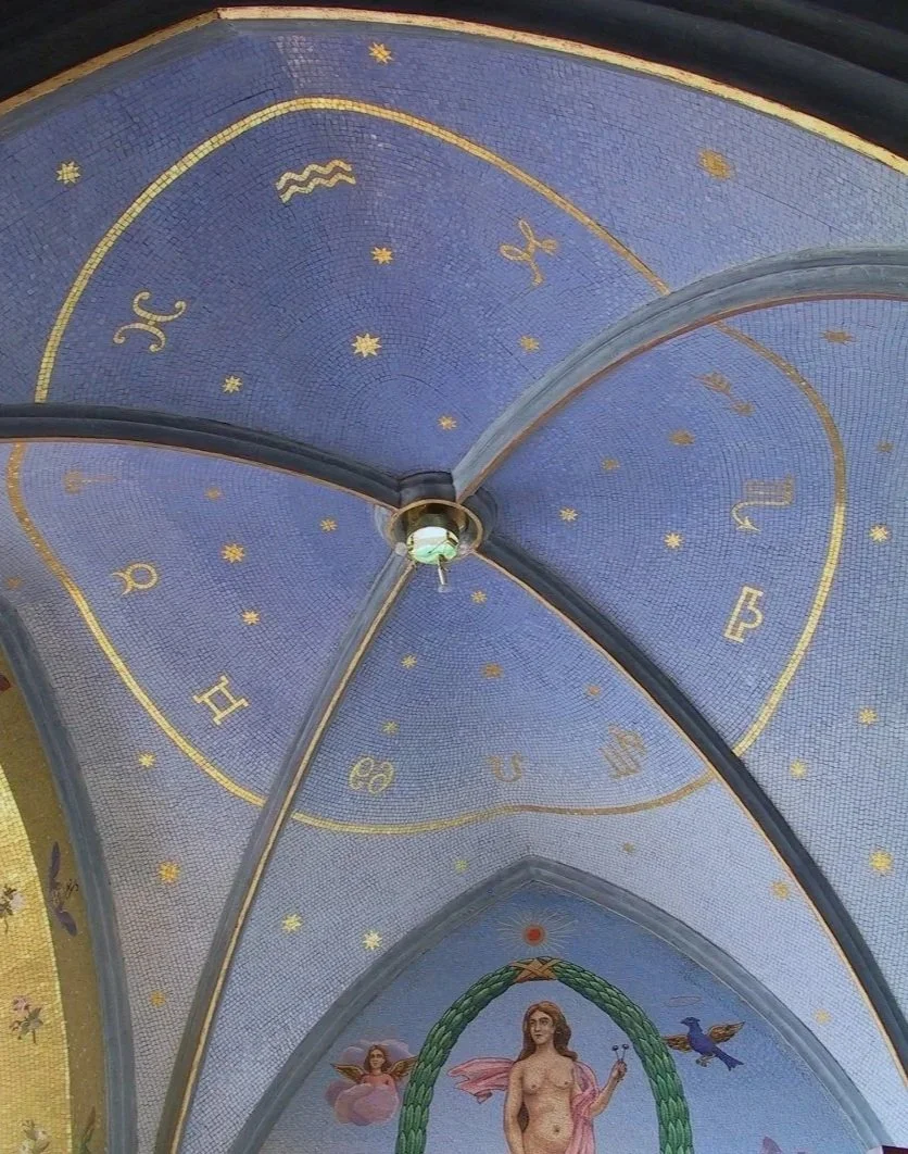 Ceiling of a church or chapel decorated with a painted constellation chart, featuring zodiac symbols and stars, with an angelic figure at the bottom surrounded by a green wreath and a bird flying nearby.
