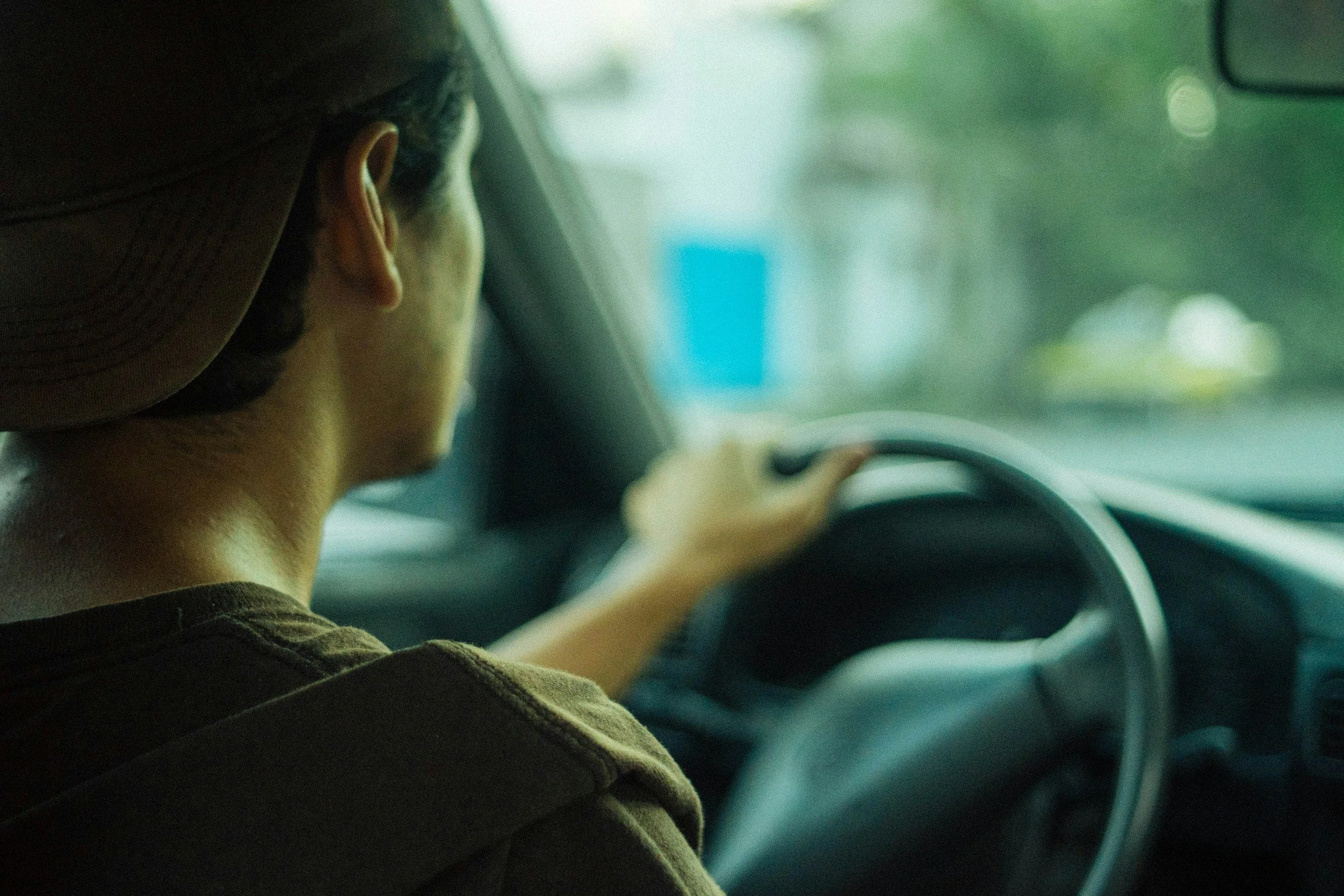 Person driving a car, viewed from behind, wearing a brown cap and a brown shirt, with a focus on their head and shoulder, with a blurred background outside the vehicle.