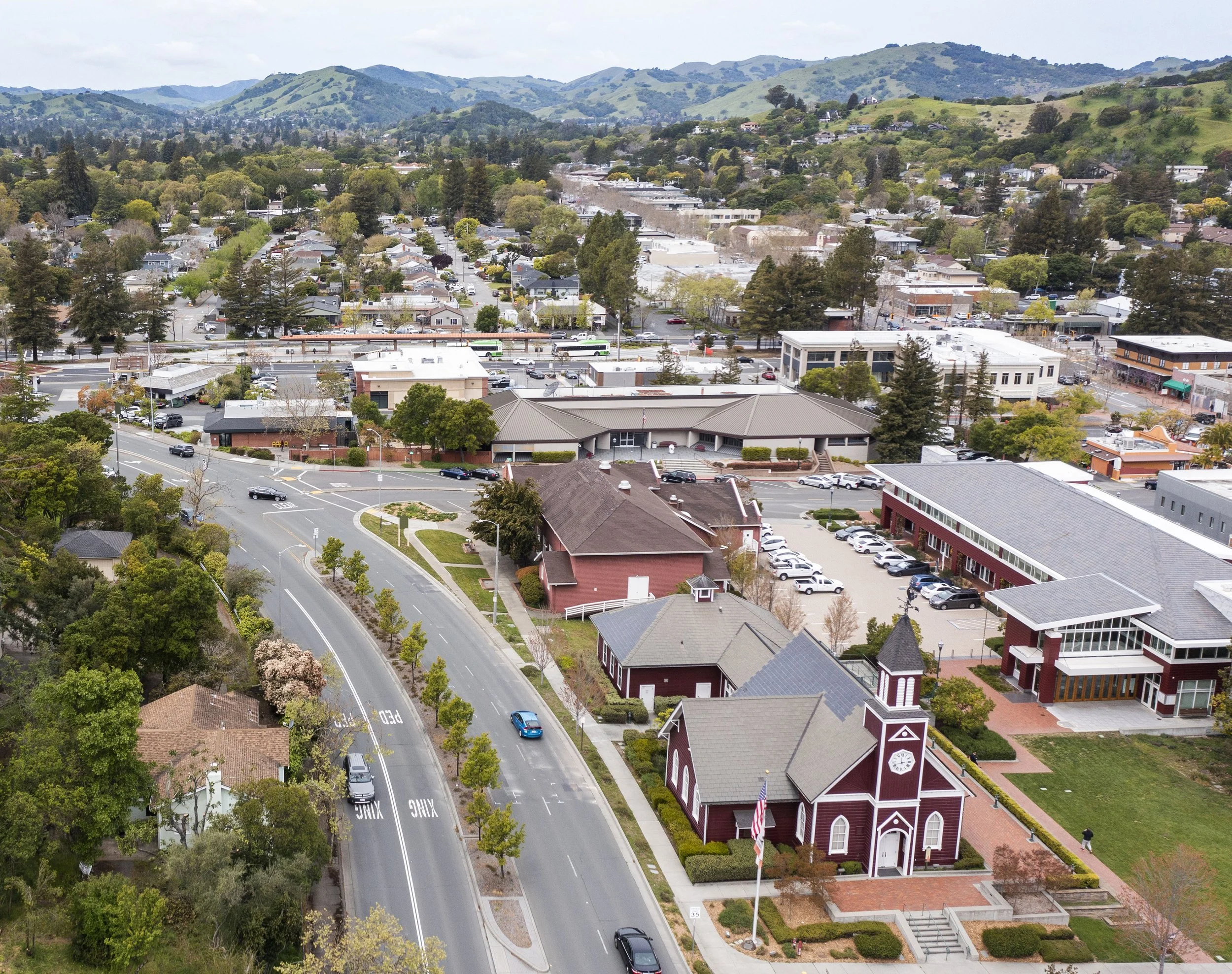 Aerial view of a small town with a church, surrounded by lush green trees and rolling hills in the background.