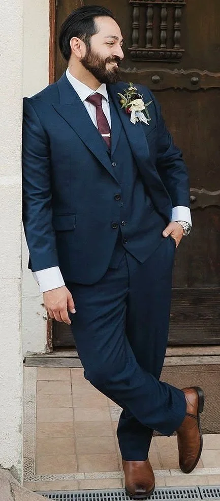 Man in a dark blue suit with a white shirt and maroon tie, smiling and leaning against a wall near a wooden door. He has a boutonniere on his left lapel, a wristwatch, and brown shoes.