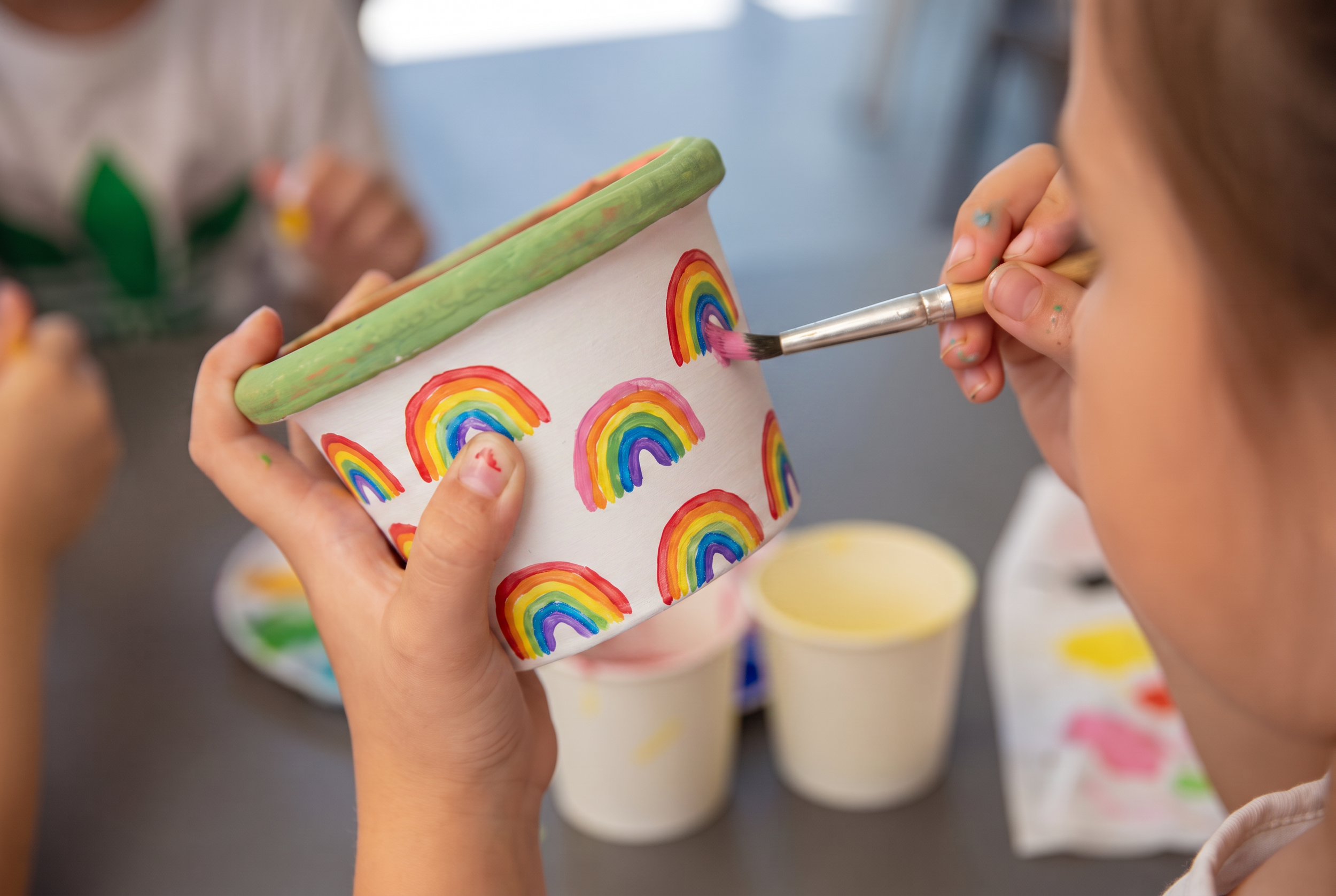 Children painting rainbows on a ceramic mug during an art activity.