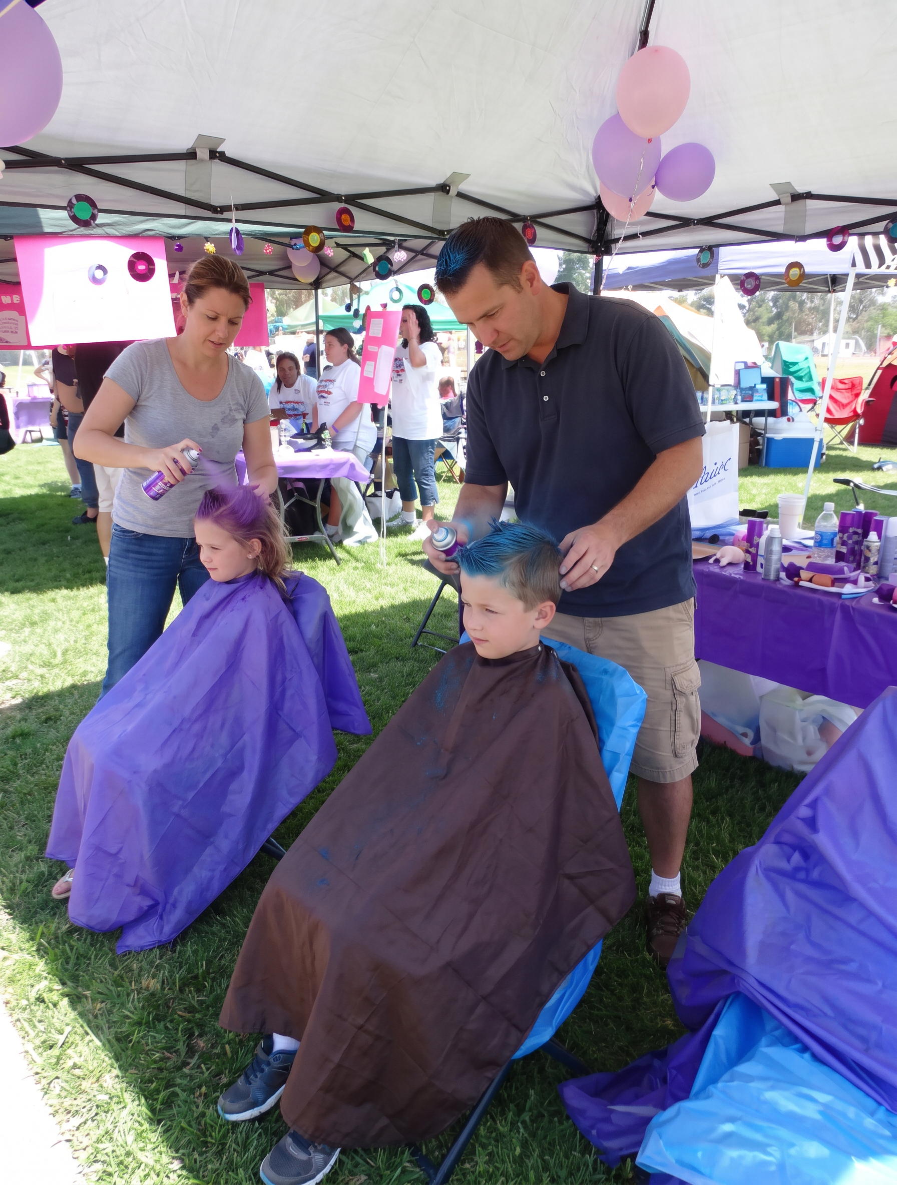 People at an outdoor event under a tent, with children getting their hair colored and decorated with balloons and decorations hanging from the tent ceiling.