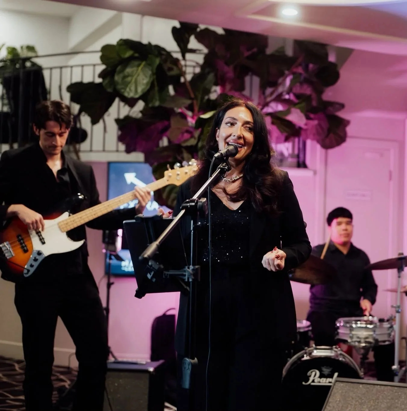 A female singer in a black dress singing into a microphone, with a bass guitarist and a drummer performing behind her, under pink stage lighting.