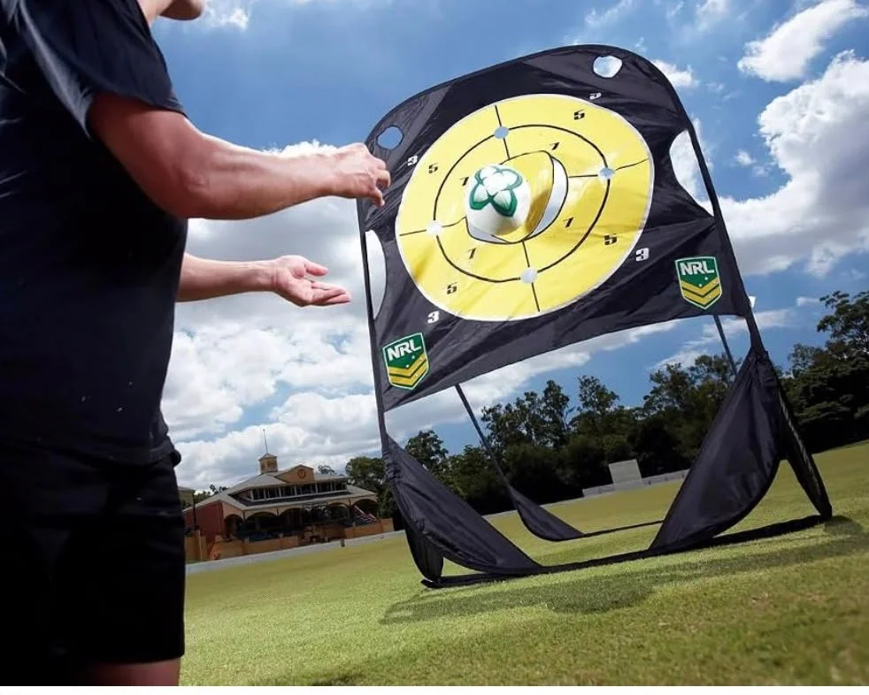 Person practicing golf with a pitching wedge next to a target mat with a yellow bullseye pattern and a foam golf ball in the air, outdoors on a grassy field under a partly cloudy sky.
