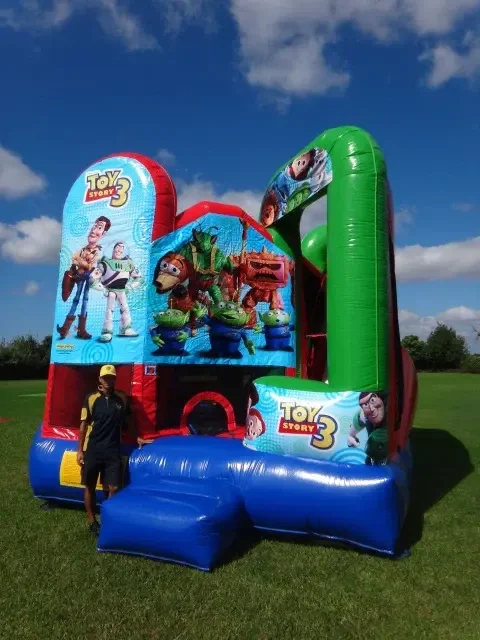 Inflatable bounce house featuring Toy Story 3 characters, with a child standing in front of it on a grassy field under a partly cloudy sky.