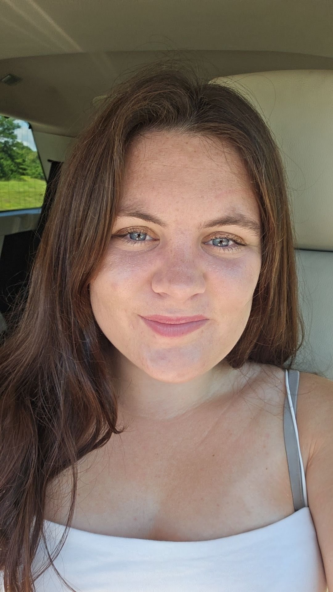 Close-up of a woman with long brown hair and blue eyes, smiling softly, sitting in a vehicle with green trees visible through the window.