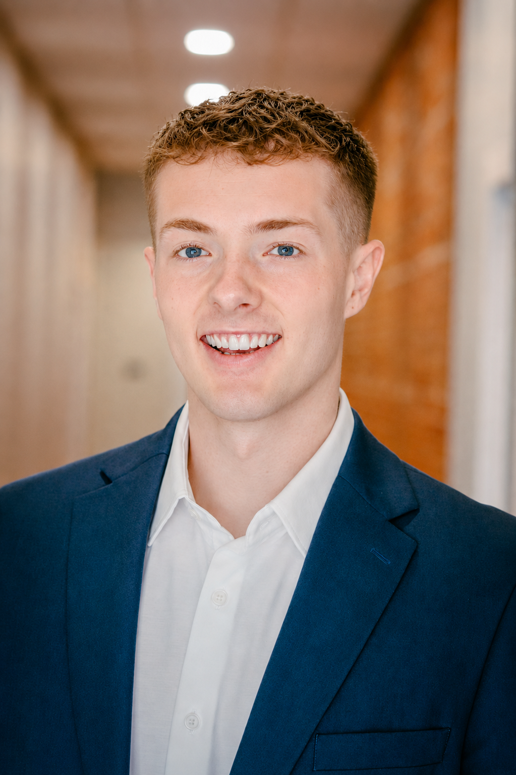 Headshot of a young man with short curly red hair and blue eyes, wearing a navy blue blazer over a white collared shirt, smiling in an indoor setting with blurred background.