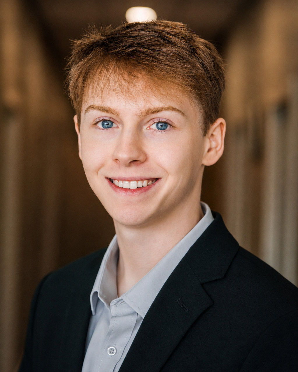 Young man with red hair and blue eyes in a suit and light gray shirt, smiling at the camera.