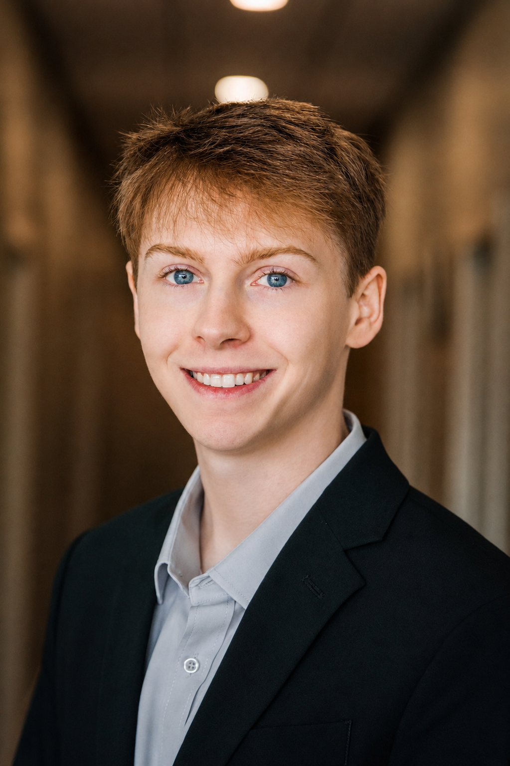 A young man with red hair and blue eyes, wearing a black suit jacket and light gray shirt, smiling in a professional headshot.