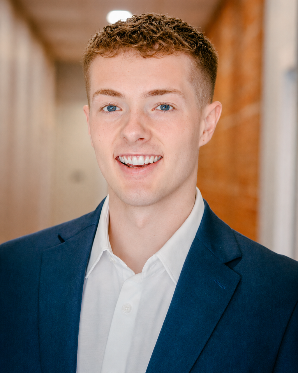 A young man with short curly hair, blue eyes, and fair skin, dressed in a navy blazer and white shirt, smiling in an indoor setting with a blurred background.