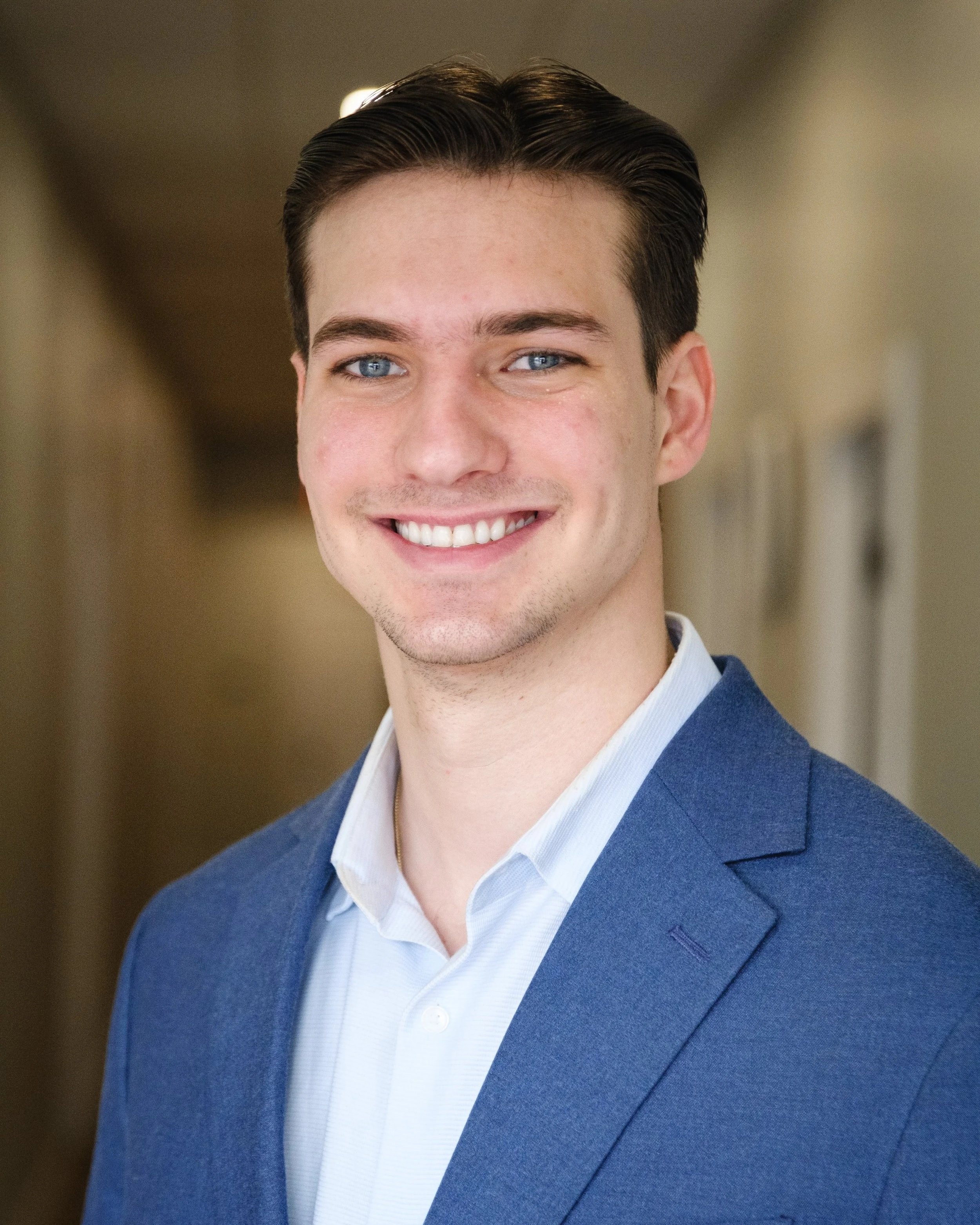 Close-up of a young man with short dark hair, blue eyes, and a bright smile, wearing a dark blue suit jacket and a light blue dress shirt, in an indoor setting.