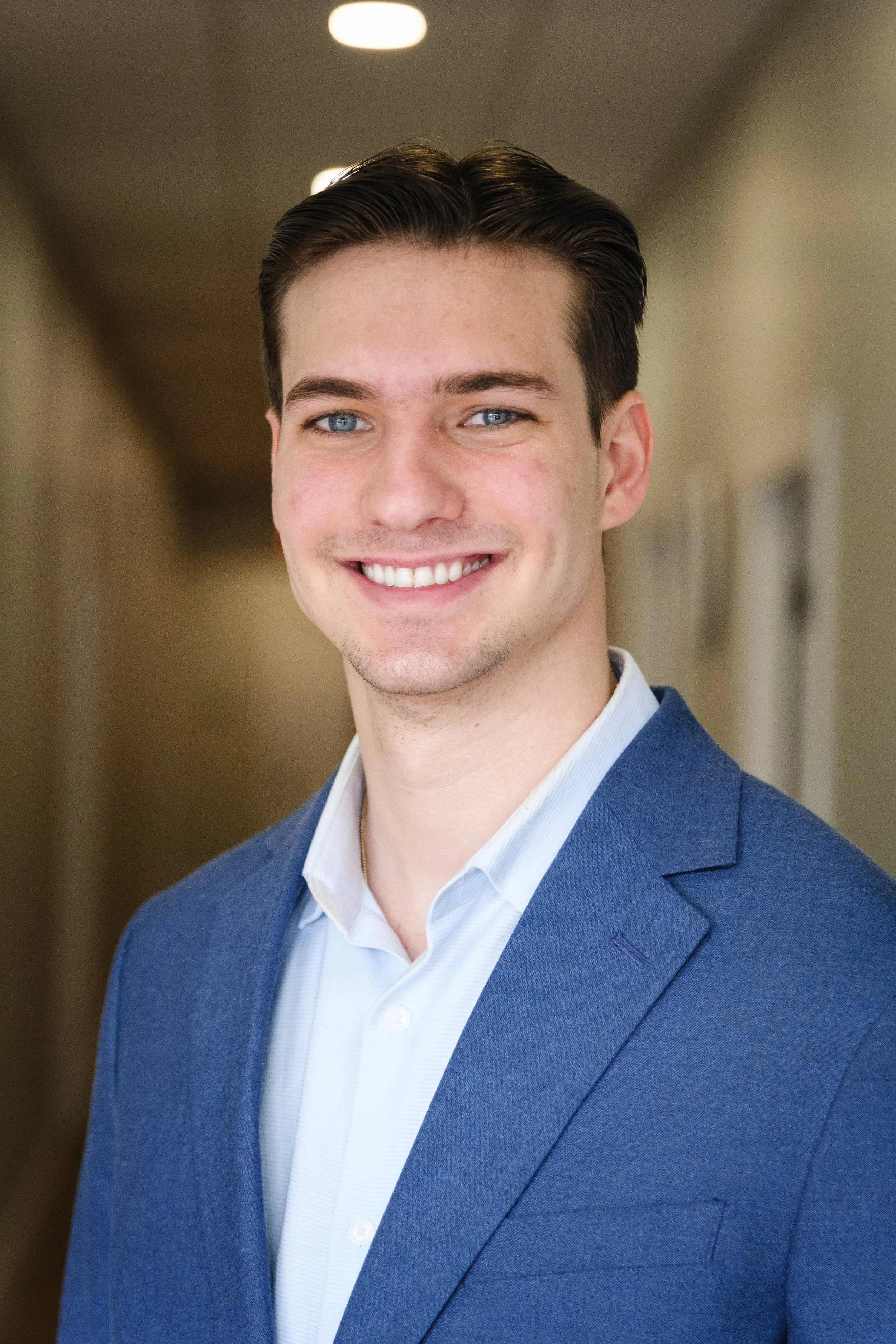 A young man with dark hair and blue eyes, dressed in a blue suit jacket and a white shirt, smiling in an indoor setting with neutral-colored walls and ceiling lights.
