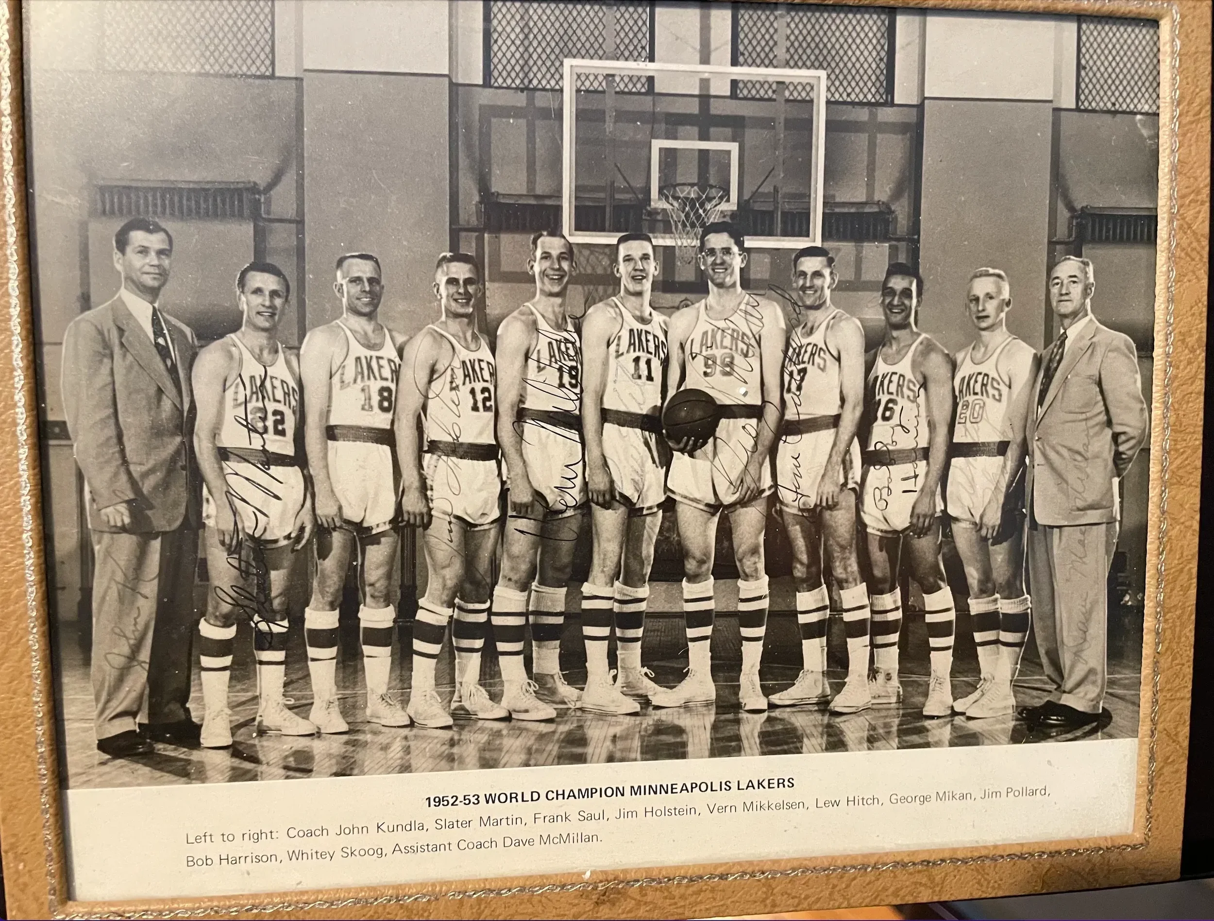 Black-and-white photograph of the 1952-53 Los Angeles Lakers basketball team, including players and coaches, standing in a gymnasium in front of a basketball hoop. The team members are dressed in Lakers uniforms, and some have autographs on the photograph. The caption mentions the team as the 1952-53 World Champion Minneapolis Lakers, with names of players and coaches listed.