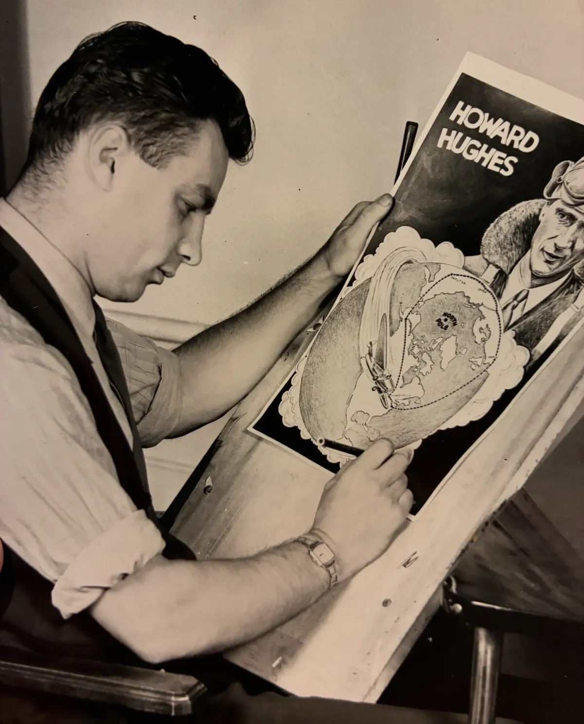 A young man with dark hair, wearing a striped shirt and a watch, is drawing a map on a piece of paper at a desk. Behind him is a poster with the name "Howard Hughes" and an illustration of Howard Hughes with goggles and a scarf.