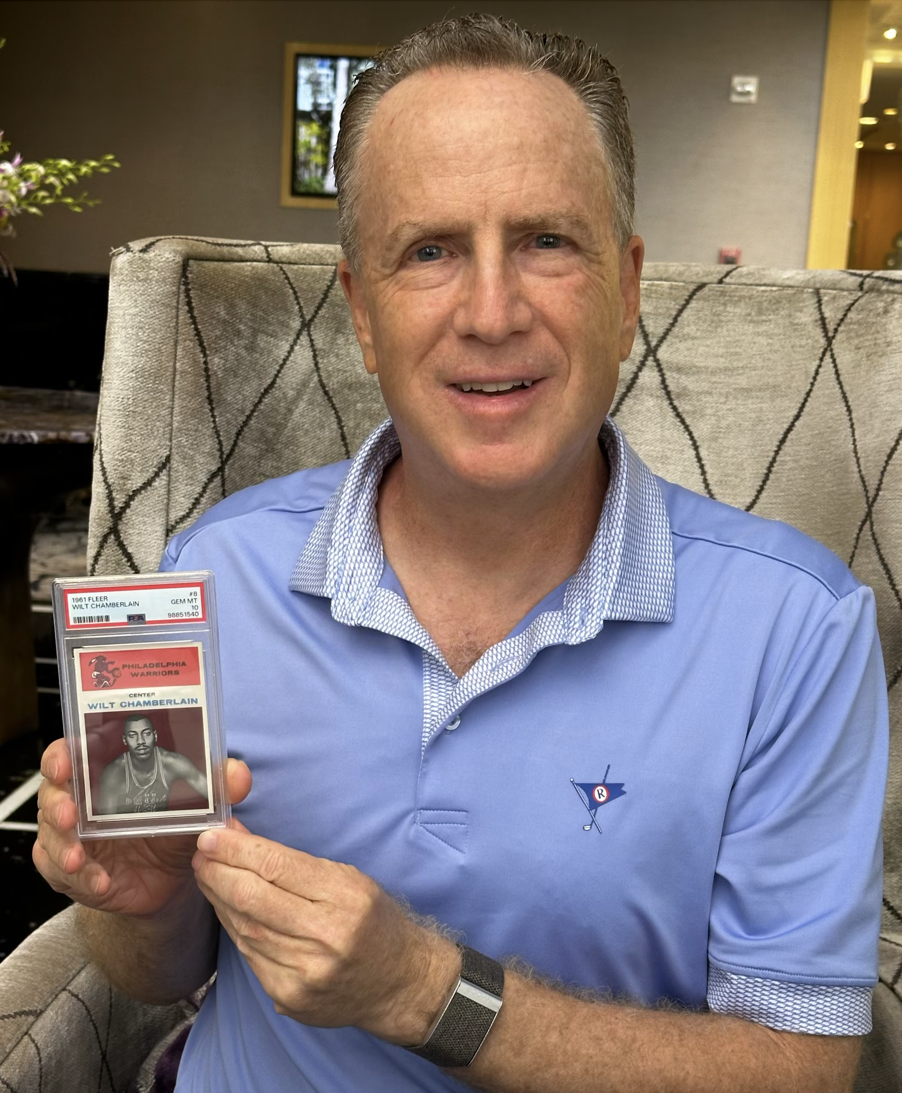 A man with short gray hair and blue eyes sitting on a beige chair with a diamond pattern, smiling and holding a black-and-white vintage basketball trading card of Wilt Chamberlain in his left hand, wearing a blue dress shirt with a small logo of a blue and red flag with a white circle and a red star on the left side of his chest. The background shows a window with a view of trees and a yellow door, with an indoor setting.