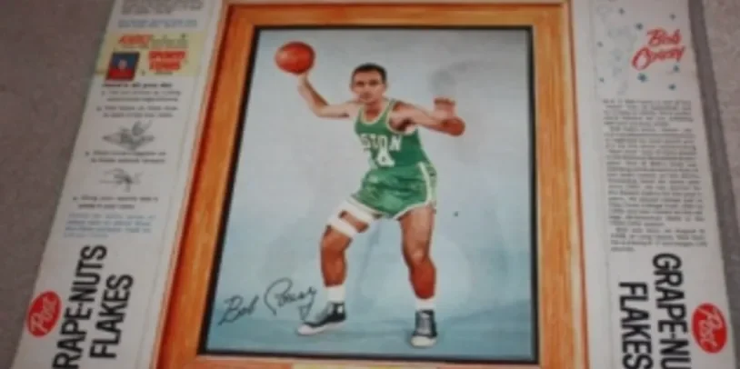 Framed photo of a basketball player in green uniform with the number 12, holding a basketball, signed by the player. The frame is on a flat surface surrounded by newspapers or advertisements.