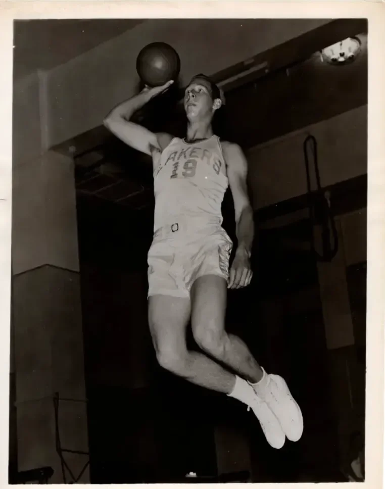 Black and white photo of a male basketball player wearing a Lakers jersey, mid-air during a basketball game, holding a basketball with one hand, with a serious expression.